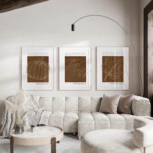 May include: Three framed art prints with botanical illustrations in brown tones. The prints feature a pumpkin, oak leaves and acorns, and a leaf, each with a Bible verse. The frames are white, and the prints are displayed above a cream-colored sofa.