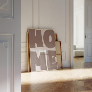 May include: A framed poster with the word "HOME" in large, brown, stylized letters. The poster is leaning against a white wall in a room with wooden floors.