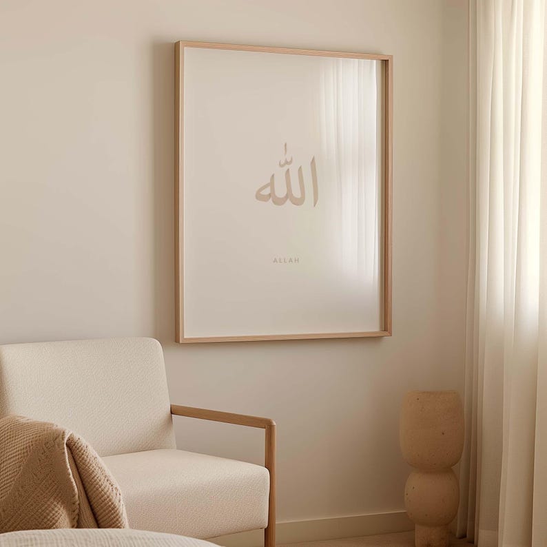 May include: A framed print featuring the Arabic word "Allah" in a light brown colour, with the word "Allah" written in English below it. The print is hanging on a light beige wall, with a white armchair and a wooden stool in the foreground.