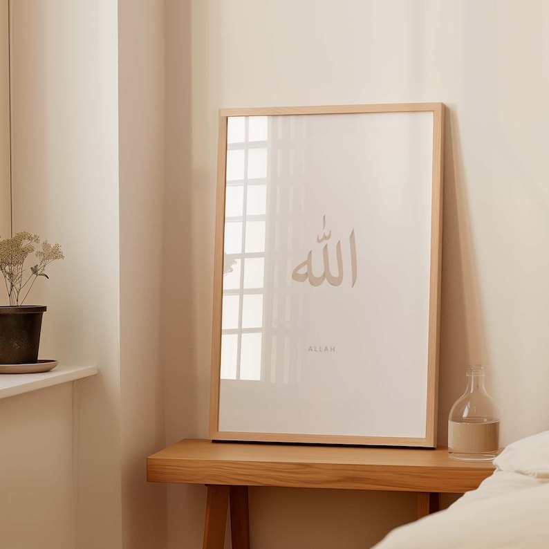 May include: A framed print featuring the Arabic word for "Allah" in a beige colour, with the word "Allah" written in English below it. The print is in a light wood frame and is hanging on a white wall.
