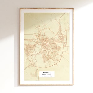 Può includere: Una stampa di mappa beige e marrone di Medina, Arabia Saudita. La mappa mostra le strade e i punti di riferimento della città. Il testo "Medina, Saudi Arabia 24°28'16.7"N 39°23'14.8"E" è stampato nella parte inferiore della mappa.