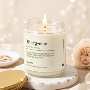 May include: A clear glass jar candle with a lit wick and a cream-colored label. The label features the text Thirty-nine, a phonetic pronunciation, and a definition describing the age before forty. The candle sits on a white marble base.