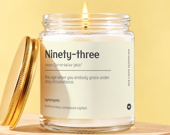 May include: A clear glass candle jar with a gold lid and a lit wick. The label features the text Ninety-three, a phonetic pronunciation, and the definition: the age when you embody grace under any circumstance, with synonyms balance boss and composure captain.