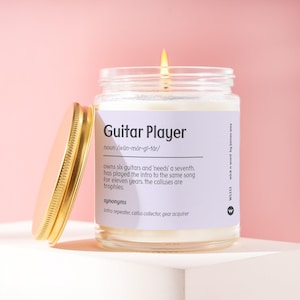 May include: A clear glass candle jar with a lit wick and a gold-colored metal lid resting beside it. The label features the text Guitar Player, noun, phonetic spelling, and a humorous definition about owning six guitars and needing a seventh.