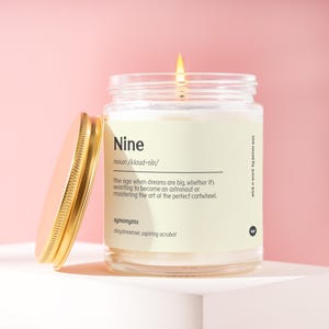May include: A clear glass jar candle with a lit wick and a gold-colored metal lid placed to the side. The label features the text Nine, noun/klaud-nin/, and a definition about big dreams, plus the synonyms daydreamer and aspiring acrobat.