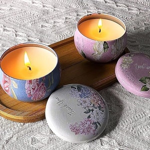 May include: Three decorative candle jars with floral designs. The jars are filled with wax and have wicks. The jars are on a wooden tray. The jars have the words "Good Luck" and "Thank You" printed on them.