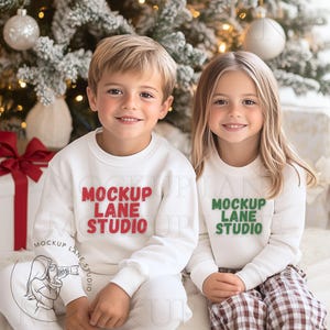 May include: Two children wearing white long-sleeved shirts with the text "Mockup Lane Studio" in red and green. The boy is wearing a white shirt with red text and the girl is wearing a white shirt with green text. They are sitting in front of a Christmas tree.