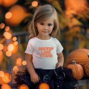 Peut inclure: Une jeune enfant porte un t-shirt blanc avec le texte "MOCKUP LANE STUDIO" en orange. L'enfant porte un tutu noir et se tient devant des citrouilles. L'arrière-plan présente des lumières oranges floues.