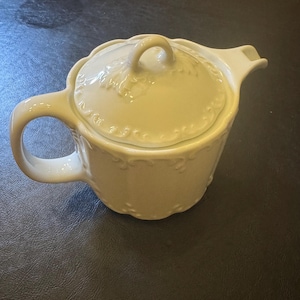 May include: A white ceramic teapot featuring a rounded body with vertical fluted panels and embossed scrollwork details. The lid includes a matching embossed pattern and a central loop handle, complemented by a curved spout and a side handle.