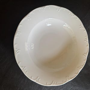 May include: A white ceramic serving bowl with a scalloped edge and embossed floral design. The bowl is round and has a deep center. A measuring tape is visible in the upper left corner.