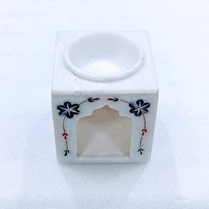 Könnte beinhalten: Ein weißer Marmor-Ölbrenner mit quadratischer Basis und einer kreisförmigen Schale oben. Die Vorderseite hat eine bogenförmige Öffnung und ist mit blauen Blumen- und Blattmotiven verziert. Der Brenner ist für die Aromatherapie konzipiert.