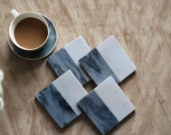 Könnte beinhalten: Vier quadratische Marmoruntersetzer mit zweifarbigem Design in Weiß und Blau-Grau. Eine weiße Teetasse mit Kaffee steht auf einer passenden Untertasse. Die Untersetzer sind auf einer hellbraunen, strukturierten Oberfläche angeordnet.
