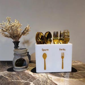 Puede incluir: Un portacubiertos blanco con letras doradas y diseños dorados de cuchara y tenedor. El soporte contiene cucharas y tenedores dorados. Un jarrón con flores secas y un quemador de aceite de piedra decorativo también están en la imagen.