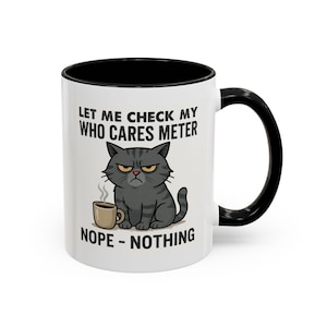 Puede incluir: Taza de cerámica blanca con interior y asa negros. La taza presenta una ilustración de un gato de dibujos animados gruñón con el texto "LET ME CHECK MY WHO CARES METER NOPE - NOTHING". Una pequeña taza de café está al lado del gato.