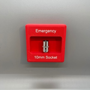 Puede incluir: Una caja de plástico roja con las palabras "Emergency" y "10mm Socket" impresas en ella. Una llave de vaso de plata está dentro de la caja.