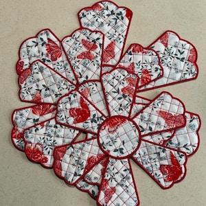 May include: A decorative, quilted floral trivet with a red and white butterfly pattern. The trivet is made of multiple petal-shaped pieces, each with a red border and a quilted design. The center is a circle, also with a red border.