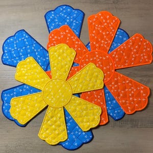May include: A colorful, quilted trivet in a flower design. The trivet features petals in blue, orange, and yellow, with white stitching and a repeating pattern of small bird footprints. The trivet is designed to protect surfaces from hot dishes.