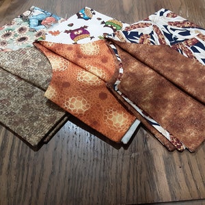 May include: Four fabric squares with different autumn-themed patterns. The squares are folded and arranged on a wooden surface. The patterns include sunflowers, owls, corn, and a brown speckled design.