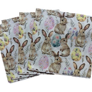 May include: A stack of Easter-themed paper napkins. Each napkin features a repeating pattern of brown bunnies, decorated Easter eggs, and floral accents on a white background. The napkins are folded and stacked.