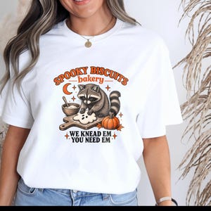 Peut inclure: Un t-shirt blanc avec un motif représentant un raton laveur cuisinant avec un biscuit en forme de fantôme, une citrouille et un bol. Le texte indique : SPOOKY BISCUITS bakery WE KNEAD EM YOU NEED EM. Le design inclut des tons orange et marron.
