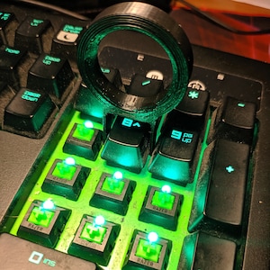 Pode incluir: Um teclado preto com teclas retroiluminadas verdes. As teclas são etiquetadas com as letras 'A', 'S', 'D', 'F', 'G', 'H', 'J', 'K', 'L', 'Z', 'X', 'C', 'V', 'B', 'N', 'M', 'Q', 'W', 'E', 'R', 'T', 'Y', 'U', 'I', 'O', 'P', '1', '2', '3', '4', '5', '6', '7', '8', '9', '0', '-', '=', '[', ']', ';', '"', ',', '.', '/', 'Backspace', 'Tab', 'Caps Lock', 'Shift', 'Enter', 'Space', 'Control', 'Alt', 'Delete', 'Insert', 'Home', 'End', 'Page Up', 'Page Down', 'Arrow Up', 'Arrow Down', 'Arrow Left', 'Arrow Right', 'Num Lock', 'Scroll Lock', 'Pause/Break', 'Print Screen', 'F1', 'F2', 'F3', 'F4', 'F5', 'F6', 'F7', 'F8', 'F9', 'F10', 'F11', 'F12'. O teclado tem um anel preto no topo.