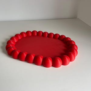 Puede incluir: Una bandeja ovalada roja con un borde elevado y texturizado. El borde está formado por una serie de pequeños bultos redondeados.