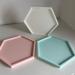 Puede incluir: Tres bandejas hexagonales en colores pastel. Una es blanca, otra rosa claro y la tercera azul claro. Las bandejas son de material sólido y tienen un borde elevado.