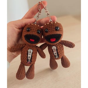 May include: Two brown crocheted doll keychains. Each doll has a zipper, black eyes, a red mouth, and small arms and legs. The keychains are attached to silver metal clasps.