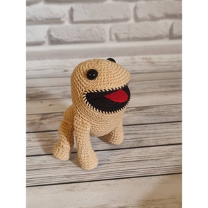 Puede incluir: Un juguete de peluche tejido a crochet con forma de monstruo marrón con ojos negros, boca negra y lengua roja. El juguete está de pie sobre una superficie de madera.