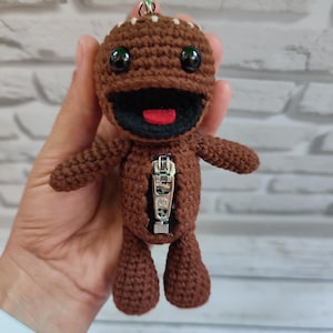 May include: A brown crocheted doll with a zipper on its torso, black eyes, and a wide open mouth with a red tongue. The doll has small arms and legs and is designed to be a keychain.