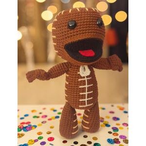 May include: A brown crocheted doll with a large head, black button eyes, and a wide mouth featuring a red tongue. The doll has cream-colored stitched details resembling a zipper down the center of the torso and along the limbs and head.