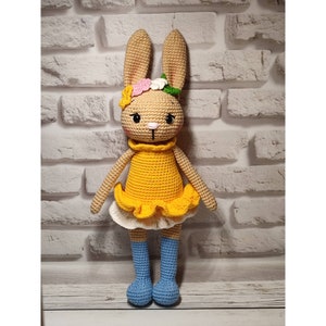 May include: A crocheted rabbit doll with tan yarn, wearing a yellow dress with a ruffled hem over a white skirt, and blue leg warmers. The rabbit has long ears, black eyes, a pink nose, and a floral crown made of pink, white, and yellow yarn.