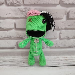 May include: A handmade green crochet zombie doll with a pink brain, black hair, and a stitched mouth. The doll has a cross stitched on its head and a white stitched design on its chest. The doll is standing on a wooden surface.