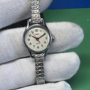 May include: A silver-tone Timex quartz wristwatch featuring a round white dial with black Arabic numerals, black hour and minute hands, and a red second hand. The watch has a flexible metal expansion band with rectangular links.