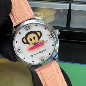 Peut inclure: Une montre-bracelet avec un boîtier en métal argenté et un bracelet en cuir texturé imitation alligator rose clair. Le cadran blanc présente le logo du singe Paul Frank, des index noirs, les chiffres 12, 3, 6 et 9, et des aiguilles argentées.