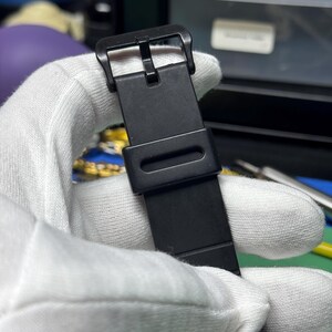 Peut inclure: Gros plan sur un bracelet de montre noir avec une boucle. Le bracelet est maintenu par une main gant&eacute;e. Le cadran de la montre est visible en bas, affichant les chiffres 12, 1 et 2. Le bracelet est en mat&eacute;riau mat.