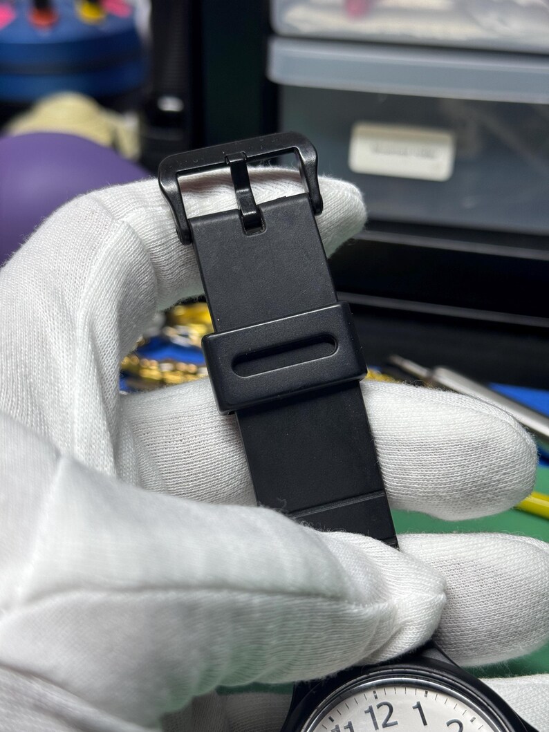 Peut inclure: Gros plan sur un bracelet de montre noir avec une boucle. Le bracelet est maintenu par une main gant&eacute;e. Le cadran de la montre est visible en bas, affichant les chiffres 12, 1 et 2. Le bracelet est en mat&eacute;riau mat.