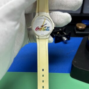 May include: A vintage cream-colored wristwatch featuring the Coca-Cola logo in red, yellow, and white on the watch face. The watch has a matching cream-colored band with several adjustment holes. The hands are yellow and the watch is held by a gloved hand.