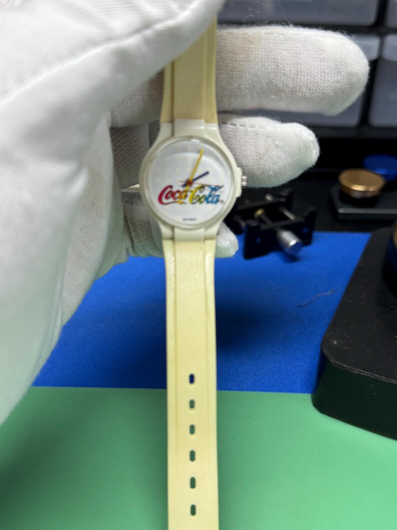 May include: A vintage cream-colored wristwatch featuring the Coca-Cola logo in red, yellow, and white on the watch face. The watch has a matching cream-colored band with several adjustment holes. The hands are yellow and the watch is held by a gloved hand.