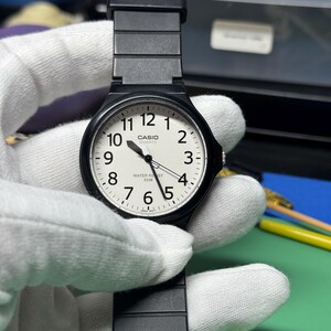 Peut inclure: Une montre Casio Quartz noire avec un cadran blanc et des chiffres noirs. La montre a un bracelet noir et est tenue par une main gant&eacute;e. Le cadran affiche l'heure et les mots "CASIO QUARTZ WATER RESIST 50M".