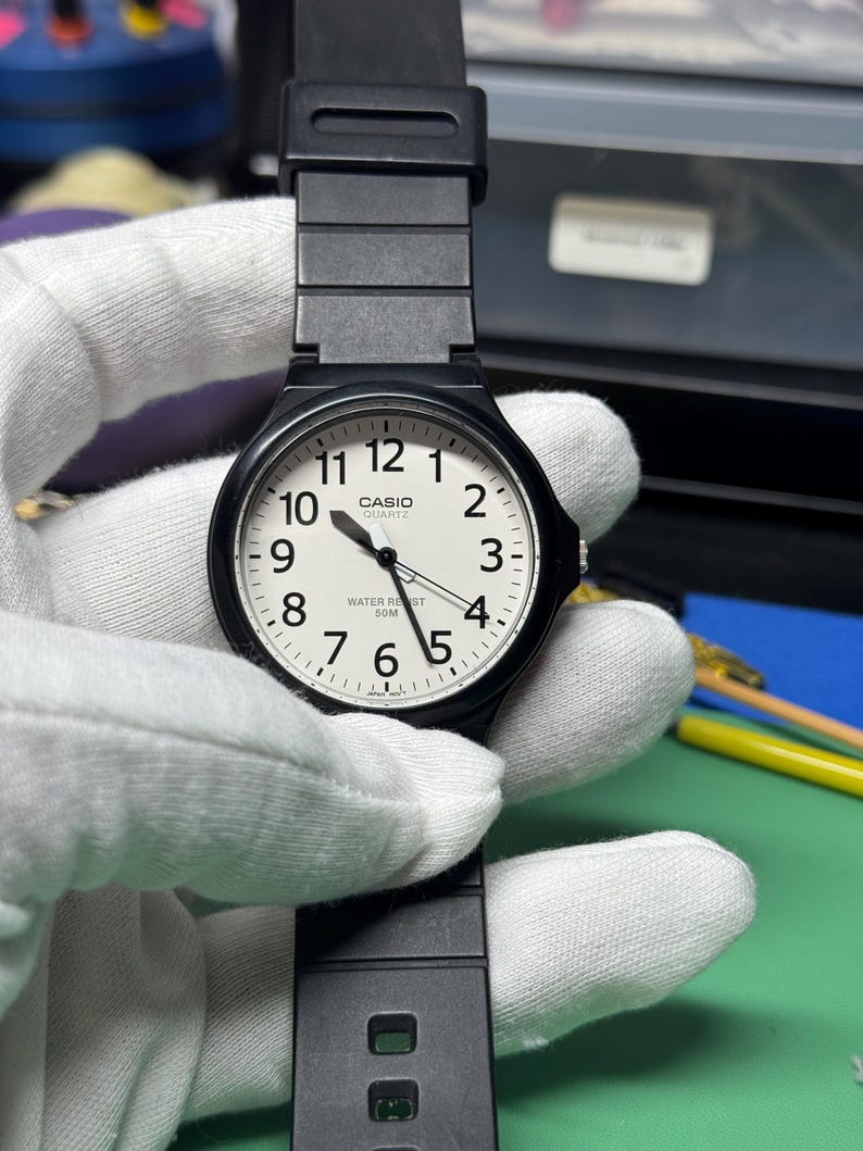 Peut inclure: Une montre Casio Quartz noire avec un cadran blanc et des chiffres noirs. La montre a un bracelet noir et est tenue par une main gant&eacute;e. Le cadran affiche l'heure et les mots "CASIO QUARTZ WATER RESIST 50M".
