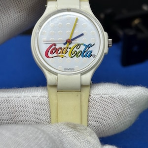 May include: A vintage Coca-Cola wristwatch with a white face and a cream-colored band. The watch face features the Coca-Cola logo in red, yellow, and blue, with a yellow second hand and blue hour and minute hands. The word "SWISS" is printed at the bottom.