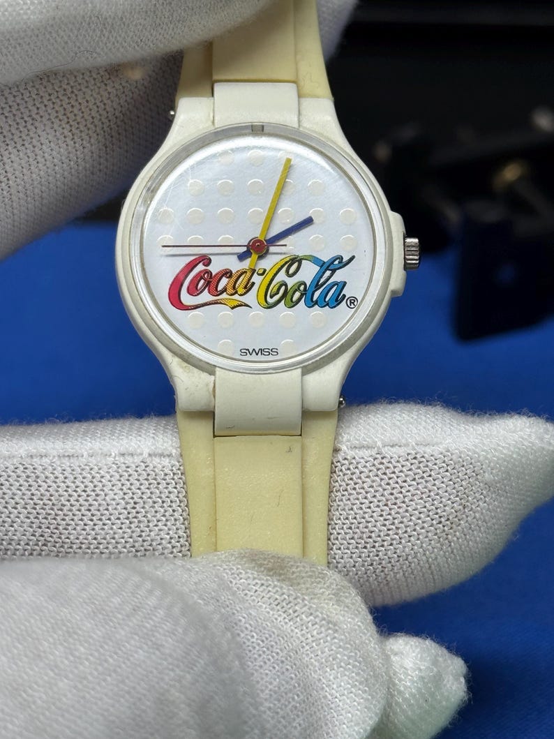 May include: A vintage Coca-Cola wristwatch with a white face and a cream-colored band. The watch face features the Coca-Cola logo in red, yellow, and blue, with a yellow second hand and blue hour and minute hands. The word "SWISS" is printed at the bottom.