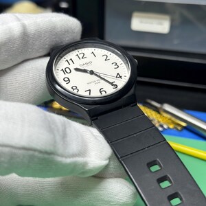 Peut inclure: Une montre &agrave; quartz Casio noire avec un cadran blanc et des aiguilles noires. La montre a un bracelet noir et est tenue par une main gant&eacute;e. Le cadran affiche le logo Casio et l'inscription "WATER RESIST 50M".