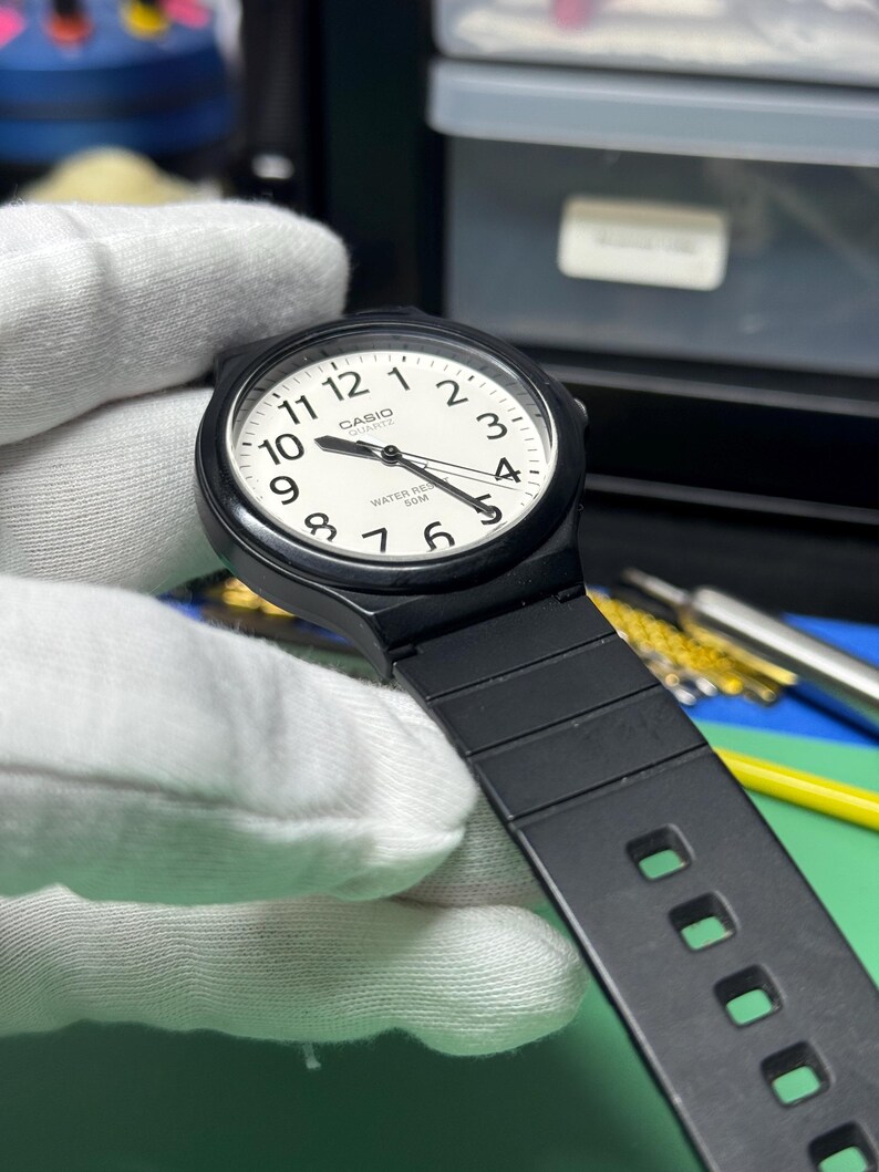 Peut inclure: Une montre &agrave; quartz Casio noire avec un cadran blanc et des aiguilles noires. La montre a un bracelet noir et est tenue par une main gant&eacute;e. Le cadran affiche le logo Casio et l'inscription "WATER RESIST 50M".