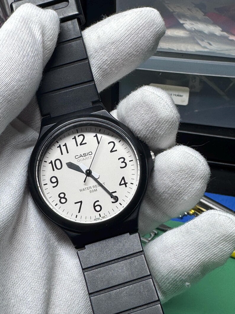 Peut inclure: Montre Casio Quartz noire avec un cadran blanc, des aiguilles noires et un bracelet noir. Le cadran affiche les chiffres de 1 &agrave; 12, avec le logo Casio et "WATER RESIST 50M" imprim&eacute; en dessous.