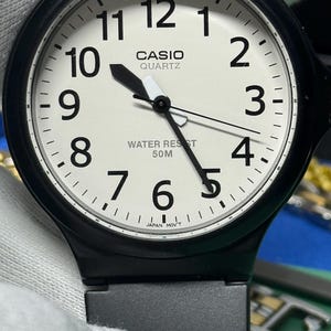 Peut inclure: Montre Casio Quartz noire avec un cadran blanc. La montre pr&eacute;sente des chiffres noirs, des aiguilles et une lunette noire. Le cadran affiche le logo Casio Quartz et "Water Resist 50M". La montre est tenue par un gant blanc.