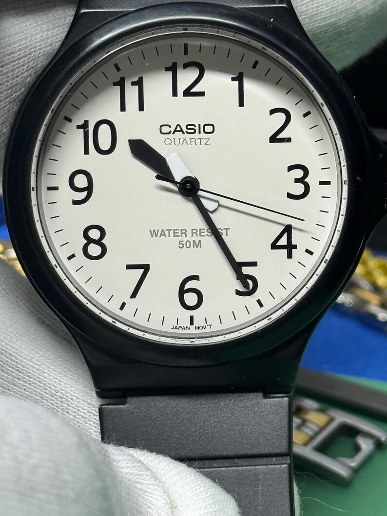 Peut inclure: Montre Casio Quartz noire avec un cadran blanc. La montre pr&eacute;sente des chiffres noirs, des aiguilles et une lunette noire. Le cadran affiche le logo Casio Quartz et "Water Resist 50M". La montre est tenue par un gant blanc.