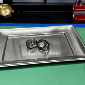 Op de afbeelding: Twee zilverkleurige ringhorloges met zwart-witte wijzerplaten liggen op een rechthoekige metalen lade. E&eacute;n horloge heeft een rechthoekige wijzerplaat, de andere een ronde wijzerplaat met een sierlijke rand. De lade staat op een groene ondergrond.