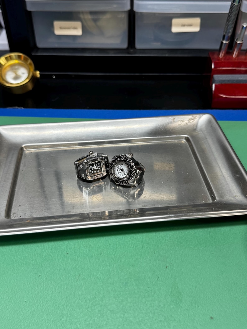 Op de afbeelding: Twee zilverkleurige ringhorloges met zwart-witte wijzerplaten liggen op een rechthoekige metalen lade. E&eacute;n horloge heeft een rechthoekige wijzerplaat, de andere een ronde wijzerplaat met een sierlijke rand. De lade staat op een groene ondergrond.